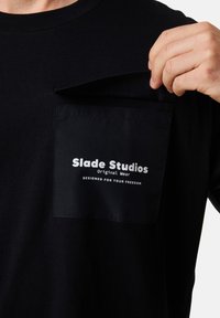 Musta t-paita, jossa on ommeltu tasku. Taskussa on valkoisella painettu teksti: "Slade Studios Original Wear SUUNNITELTU VAPAUTTAASI VARTEN."