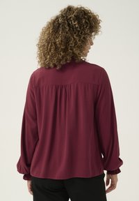 Burgundy long-sleeve blouse with gathered back yoke and elastic cuffs, made of smooth fabric. Back view, no visible patterns or hardware.
