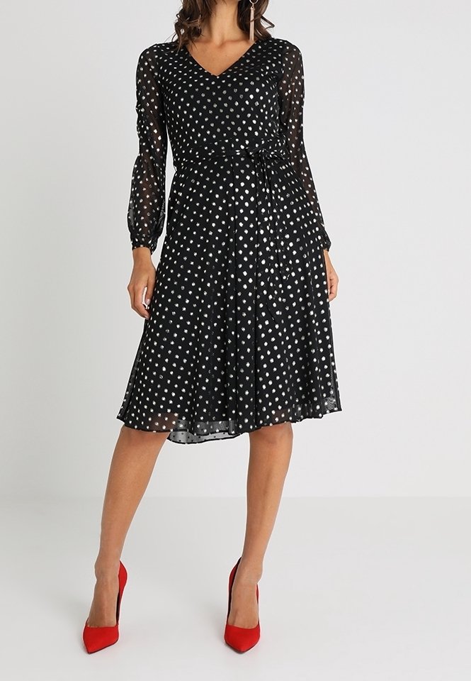 Robe noire à pois blancs avec manches longues en voile transparent, décolleté en V, taille cintrée et jupe évasée. Associée à des chaussures à talons hauts rouges.