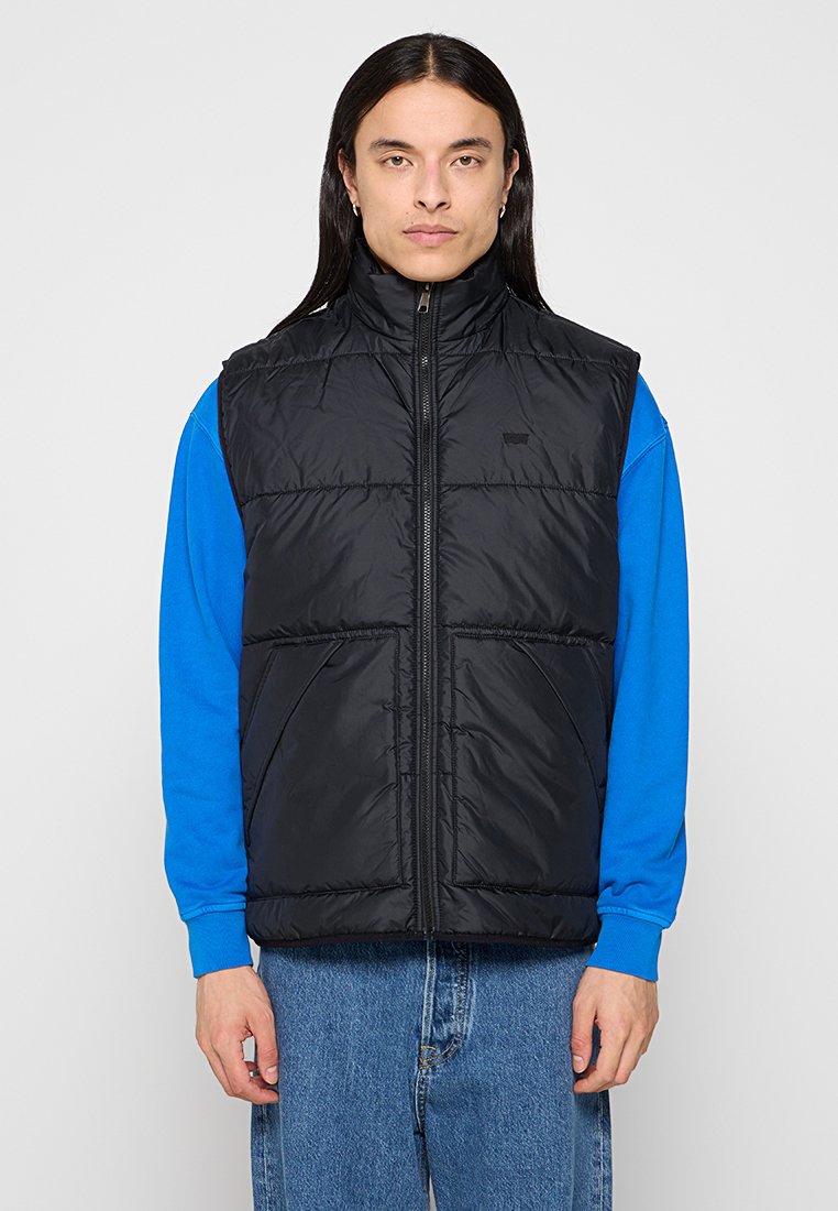 Schwarze Puffer-Weste mit hohem Kragen, Reißverschluss vorne und zwei Vorderaschen. Getragen über einem blauen Langarmshirt, vor einem weißen Hintergrund präsentiert.
