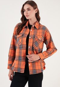 Narancssárga kockás flanel ing fekete és kék részletekkel, klasszikus gallérral, gombos záródással és két mellezsebből áll.