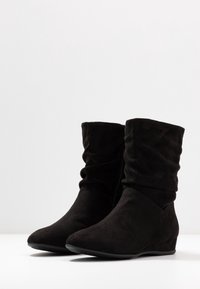 Černé kotníkové boty z broušené kůže s volným tvarem, špičatou špičkou a nízkým klínovým podpatkem. Mají boční zip pro snadné obouvání.