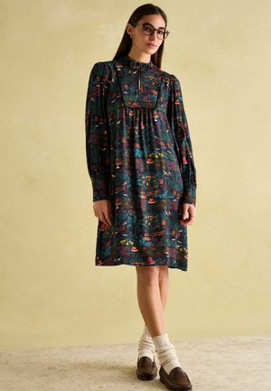 Dunkelblumenmuster Kleid mit hohem Kragen, langen Ärmeln und gerafftem Oberteil. Das Model trägt braune Slipper und cremefarbene Socken.