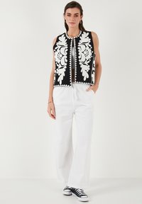 Zwarte cropped top met witte bloemenborduursels en pomponaccenten, gecombineerd met losse witte broek en zwarte sneakers.