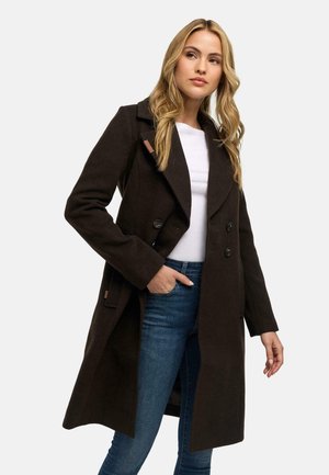 Femme blonde portant un manteau croisé marron foncé par-dessus une chemise blanche et un jean bleu, avec une main dans sa poche.
