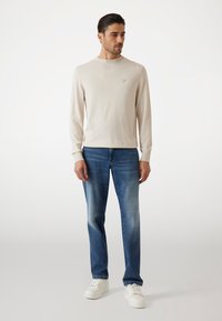 Maglietta a maniche lunghe color beige chiaro con scollatura rotonda, abbinata a jeans in denim blu medio e sneakers bianche, con un logo sottile sulla maglietta.