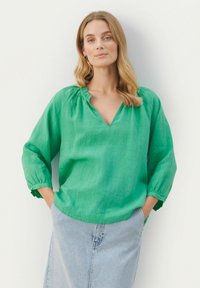 Grüne Leinenbluse mit V-Ausschnitt, gerafftem Halsauschnitt und Dreiviertelärmeln, kombiniert mit einem hellen Denimrock. Der Stoff wirkt leicht.