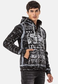 Schwarze Zip-Hoodie mit grafischen Prints, einem Totenkopfdesign und weißen Akzenten. Hergestellt aus weichem Stoff mit Kapuze und Kordelzug sowie Fronttaschen.