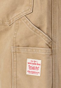 Beige canvas fabric with prominent stitching, featuring a patch with red text and a graphic. Includes a front pocket with a flap.
