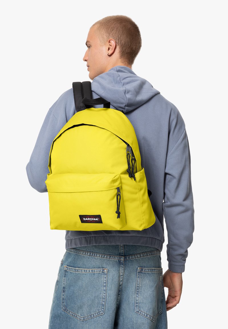 Mochila amarilla con forma redondeada, hecha de material duradero. Cuenta con dos bolsillos frontales con cremallera, correas negras y un parche con el logo.
