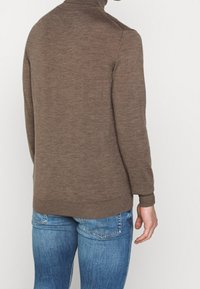 Brauner Wollpullover mit hohem Kragen und langen Ärmeln, mit glatter Textur und tailliertem Schnitt, getragen zu blauen Jeans.