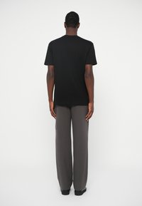 Homem de costas, vestindo uma t-shirt preta de manga curta simples e calças cinzentas largas, contra um fundo branco liso.