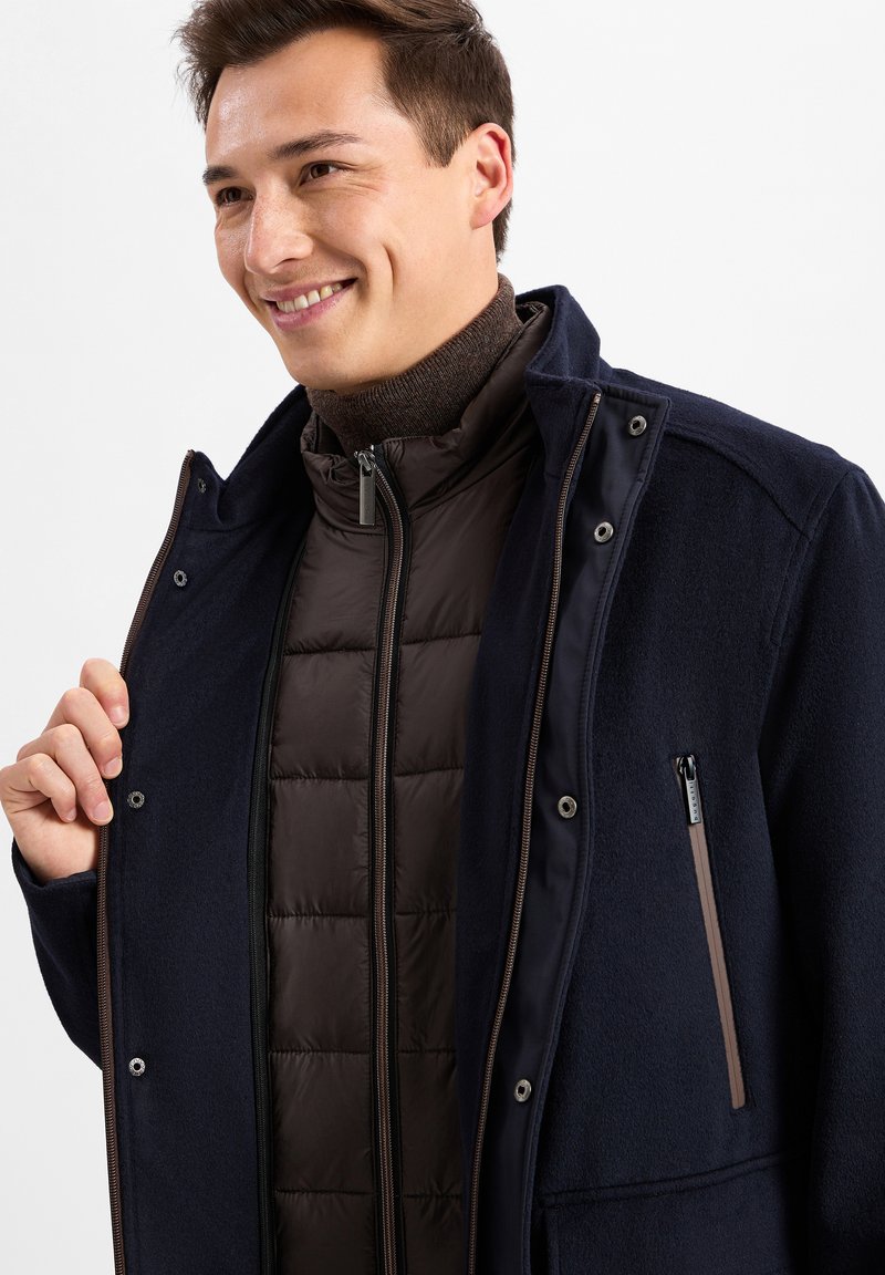 Marineblaue Außenjacke mit weicher Textur, die mit Knöpfen und Reißverschlüssen geschlossen wird. Braune isolierte Innenjacke mit gesteppter Musterung und hohem Kragen.