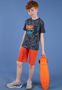 Garçon aux cheveux roux portant un t-shirt noir à motif de dinosaure et un short orange, tenant un skateboard orange sur un fond bleu.