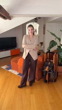 Νέα γυναίκα στέκεται με σταυρωμένα χέρια σε ένα σαλόνι δίπλα σε μια κωπηλατική μηχανή, πορτοκαλί καναπέ, φυτό και μονάδα τηλεόρασης.