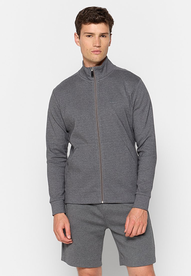 Jonge man in een grijze zip-up jas en bijpassende grijze shorts staat tegen een effen lichte achtergrond.