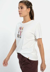 Hvid kortærmet t-shirt med et multifarvet "NATURE" grafisk motiv, parret med bordeaux bukser og en tekstureret stofoverflade.