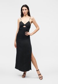 Femme debout, main sur la hanche, portant une robe noire sans manches avec des fines bretelles, une découpe à l'avant, une fente latérale et des sandales noires à talons hauts.