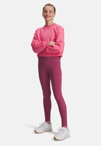 Tonårstjej står med armarna i kors, iklädd en rosa sweatshirt, lila Under Armour leggings och vita sneakers mot vit bakgrund.