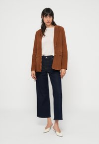 Blazer en daim marron porté sur un pull léger, associé à un jean en denim foncé taille haute et des mules crème. Coupe droite avec fermeture à un bouton.