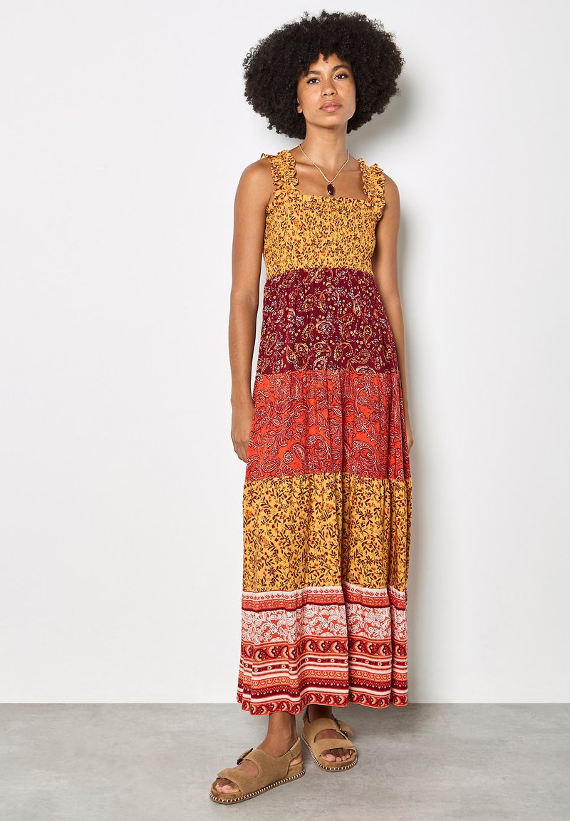 Femme aux cheveux bouclés portant une robe longue sans manches à volants multicolore avec des motifs floraux et paisley, ainsi que des sandales beige.