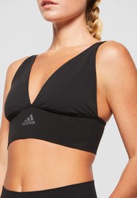Črn športni nedrček z globokim V-izrezom, širokimi naramnicami in diskretnim adidas logotipom na traku. Gladek, elastičen material.