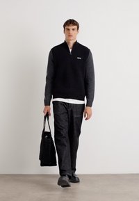 Colete com fecho éclair em preto e cinza, feito de um tecido texturizado, usado sobre uma camisa branca. O modelo segura uma mala tote preta e usa calças e sapatos escuros.