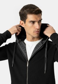 Črno jopa z zadrgo in sivo notranjostjo, na kapucni in vrvicah z vzorcem logotipa, izdelana iz udobnega blaga.