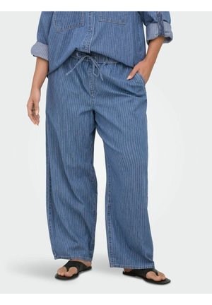 Osoba ubrana w luźne niebieskie spodnie w prążki i dopasowaną koszulę, stojąca z jedną ręką w kieszeni i czarnymi sandałami.