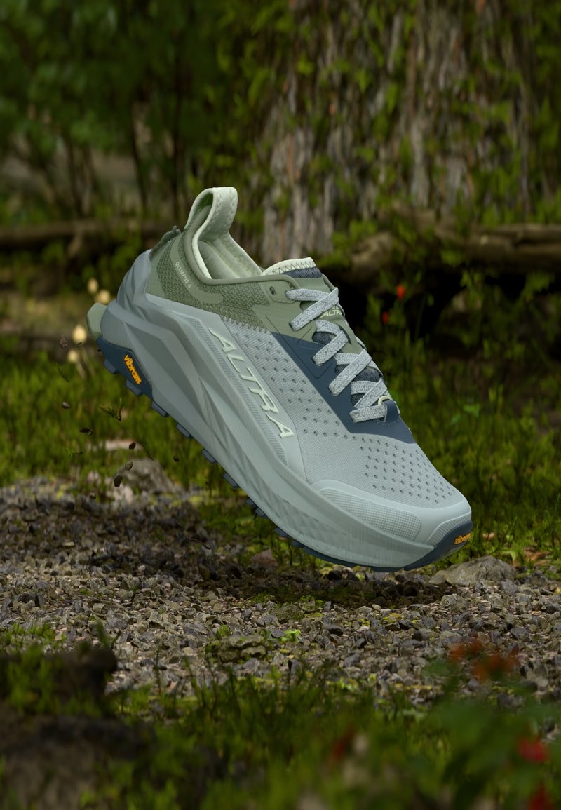 Chaussure de trail running vert clair et grise avec le logo "ALTRA", flottant au-dessus d'un sentier de gravier dans un cadre forestier avec des arbres et du feuillage.