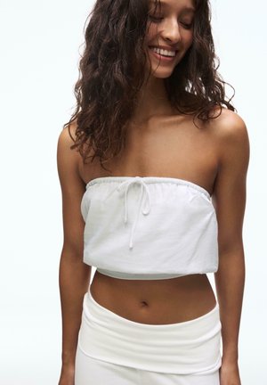 Nasmejana ženska z kodrastimi lasmi, oblečena v belo top brez naramnic in ujemajočo belo krilo na preprostem belem ozadju.