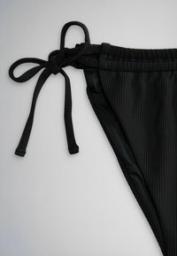 Bas de bikini noir côtelé avec des liens noués sur les côtés sur un fond clair uni.
