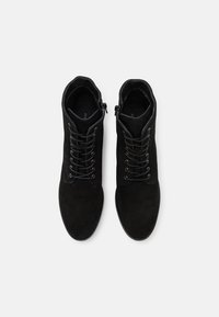 Botas de tornozelo em camurça preta com uma textura suave, apresentando atacadores e um zíper na lateral, proporcionando uma silhueta justa.