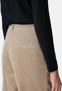 Beige corduroy trousers with vertical ribbed texture, featuring a back pocket and a snug fit, paired with a black top partially visible.