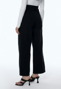 Zwarte wijde pantalon gemaakt van een gladde stof, met een hoge taille en zichtbare stiksels. Gepaarde met zwarte sandalen met hakken en bandjes.
