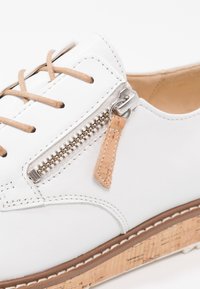 Chaussure blanche à lacets avec lacets couleur marron clair, fermeture éclair latérale métallique avec tirette marron clair, et semelle en liège texturée ornée de détails de couture blanche.