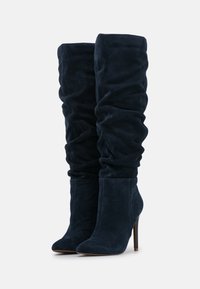Bottes en daim bleu marine montant au-dessus du genou avec un style décontracté, bout pointu et talon fin et haut. La finition texturée ajoute de la profondeur.