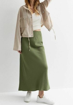 Femme portant une veste courte beige, un débardeur blanc, une jupe longue vert olive et des baskets blanches, debout devant un fond uni.