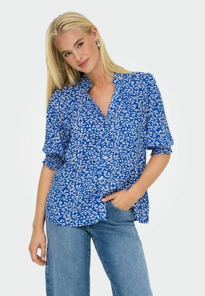 Blusa a fantasia floreale blu con maniche corte a sbuffo e colletto, realizzata in tessuto leggero. Abbinata a jeans in denim.