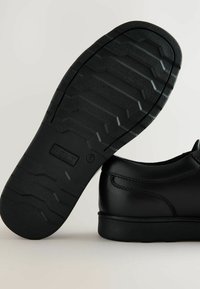 Chaussures noires en cuir avec une semelle en caoutchouc texturé présentant un motif en zigzag et un logo en relief. Le design comprend un bout arrondi et des coutures minimales.