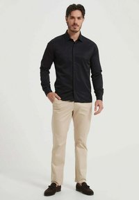 Chemise noire à manches longues avec des boutons sur le devant, accompagnée d'un pantalon beige clair et de mocassins foncés. Tissu lisse, coupe ajustée, détails minimaux.