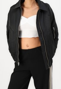 Osoba ubrana w czarną kurtkę z zamkiem błyskawicznym na białą krótką bluzkę i czarne spodnie, z rękami w kieszeniach kurtki.