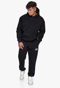 Sort hættetrøje med en frontlomme og matchende sweatpants. Bomuldsstof med en blød tekstur. Har hvidt trykt logo på bukserne. Hvide sneakers.