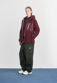Casaco de fleece cor vinho com detalhes azuis, com bolsos com fecho, combinado com calças cargo verdes escuras e ténis brancos. Estilo casual, com um ajuste descontraído.