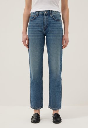 Femme portant un jean bleu coupe droite, un haut blanc rentré, et des mocassins noirs, debout devant un fond beige uni.