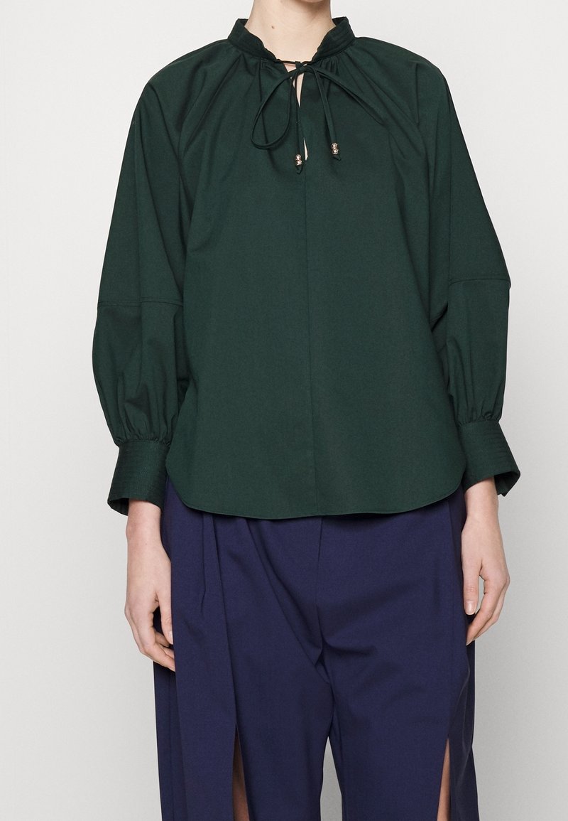 Personne portant un chemisier à manches longues vert foncé avec un col noué et un pantalon ample bleu profond fendu à l'avant, debout devant un fond uni.