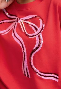 Červená mikina s potiskem mašle z flitrů v růžové, světle růžové a vínové barvě, s plynulými liniemi a texturovaným, reflexním povrchem.