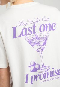 Valkoinen puuvillapaita, jossa on suuri violetti grafiikka, joka esittää martini-lasia oliiveilla ja teksti "Iso ilta ulkona, tämä on viimeinen kerta, lupaan."