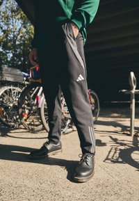 Pantalones deportivos negros con rayas laterales grises y logo de Adidas, combinados con zapatos negros de suela texturizada, en un entorno al aire libre.
