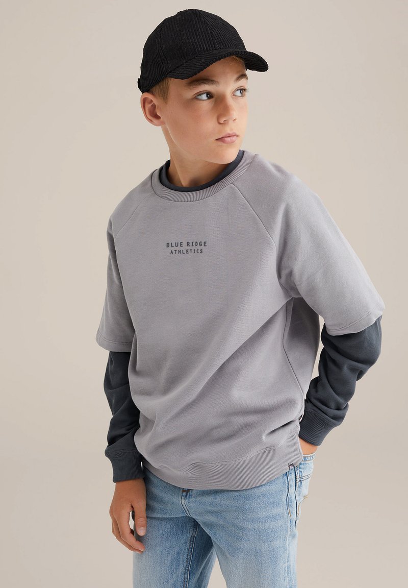 Felpa grigia con maniche corte a raglan, con la scritta "BLUE RIDGE ATHLETICS". Indossata sopra una maglietta a maniche lunghe blu navy, abbinata a jeans azzurri.